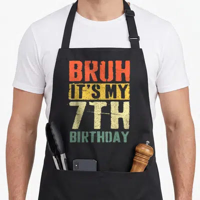 7yr birthday apron - Distressed stacked text BRUH IT'S MY 7TH BIRTHDAY in orange yellow cream and green — shown on a black cotton canvas apron against a white background