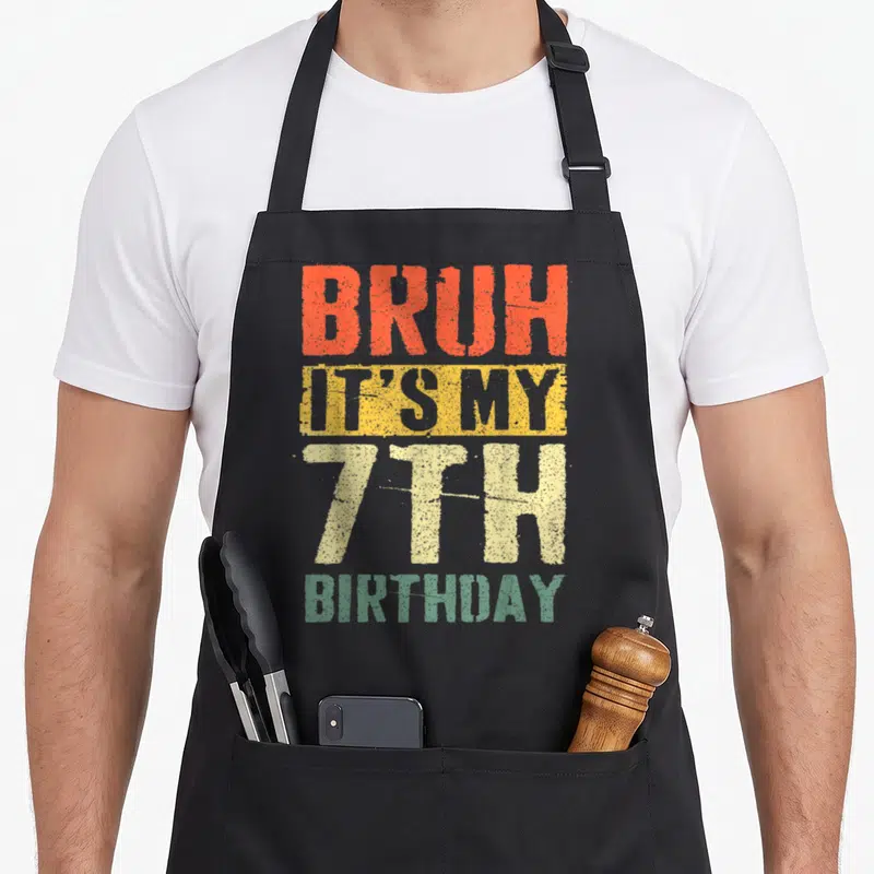 7yr birthday apron - Distressed stacked text BRUH IT'S MY 7TH BIRTHDAY in orange yellow cream and green — shown on a black cotton canvas apron against a white background