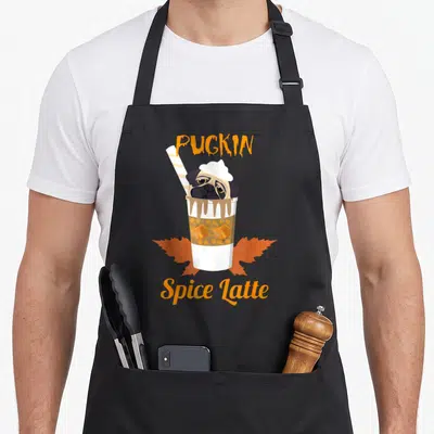 Pugkin dog apron - Cartoon pug in coffee cup with PUGKIN Spice Latte text and orange autumn leaves — shown on a black cotton canvas apron against a white background