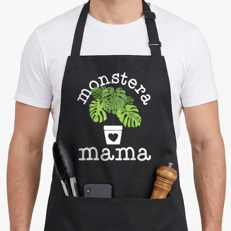 Monstera mama apron - Green monstera plant in white pot with heart and arched text — shown on a black cotton canvas apron against a white background