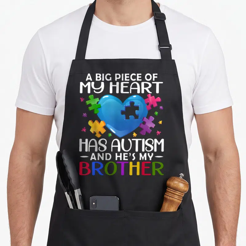 Autism brothers apron - Blue heart with puzzle cutout surrounded by multicolored pieces and supportive text — shown on a black cotton canvas apron against a white background