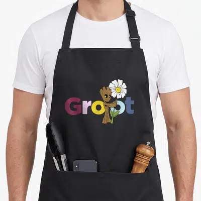 Flower galaxy apron - Cartoon Groot holding white flower in front of red yellow and blue Groot text — shown on a black cotton canvas apron against a white background