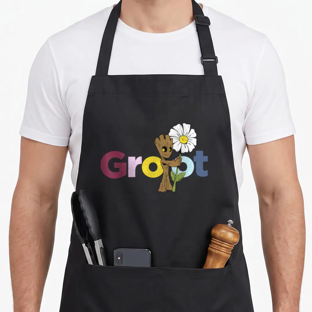 Flower galaxy apron - Cartoon Groot holding white flower in front of red yellow and blue Groot text — shown on a black cotton canvas apron against a white background