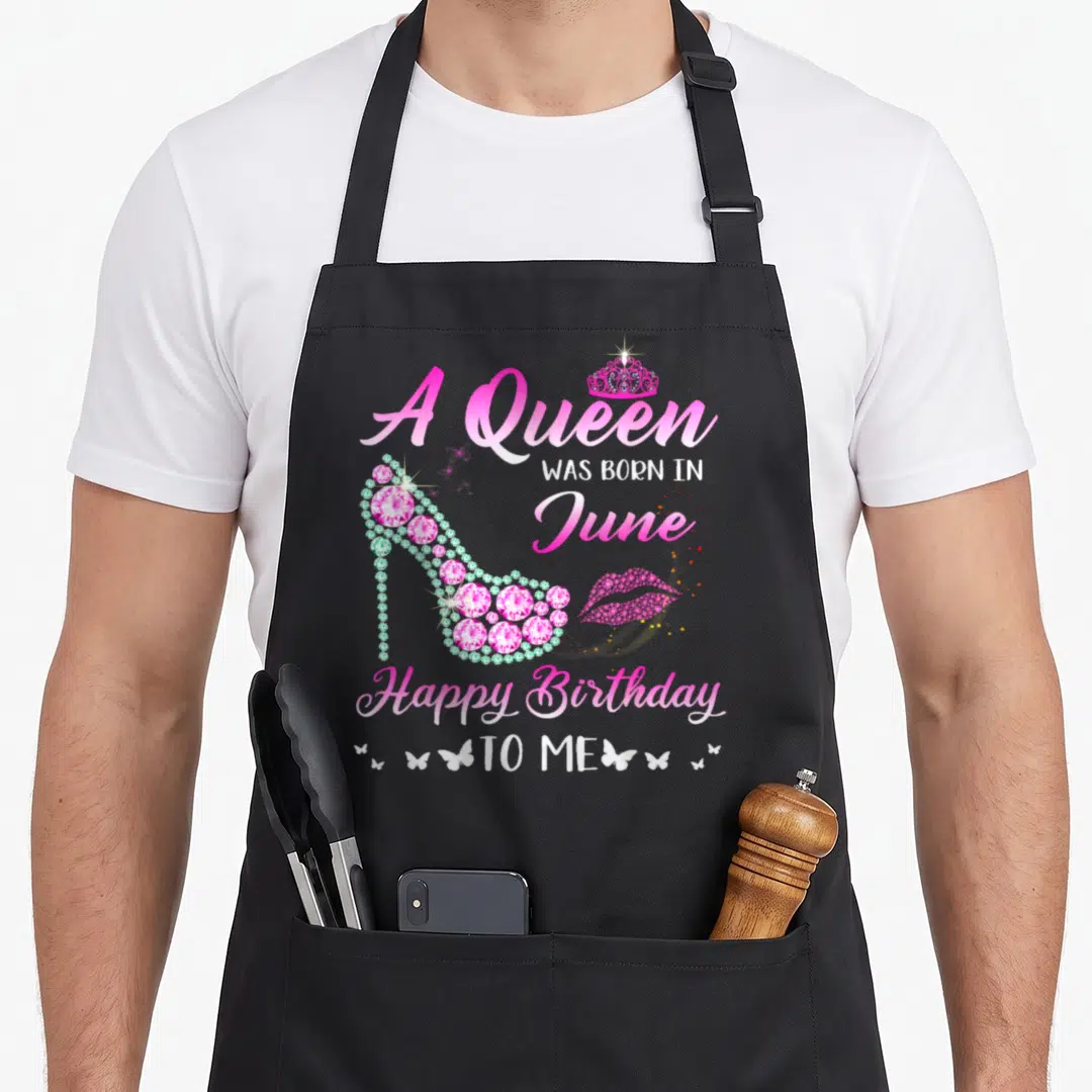 Funny june birthday apron - Jeweled pink high heel shoe with A Queen WAS BORN IN June text and crown — shown on a black cotton canvas apron against a white background