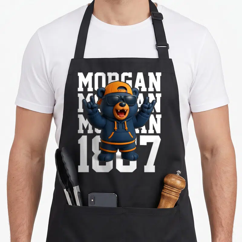 Gan apron or apron - Cartoon bear in blue hoodie and orange cap standing before stacked white MORGAN text and 1887 — shown on a black cotton canvas apron against a white background