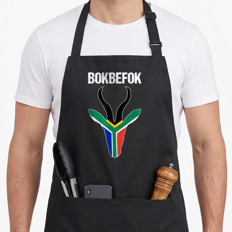 Bokbefok south african apron - White BOKBEFOK text above a springbok head filled with green, yellow, blue, red — shown on a black cotton canvas apron against a white background
