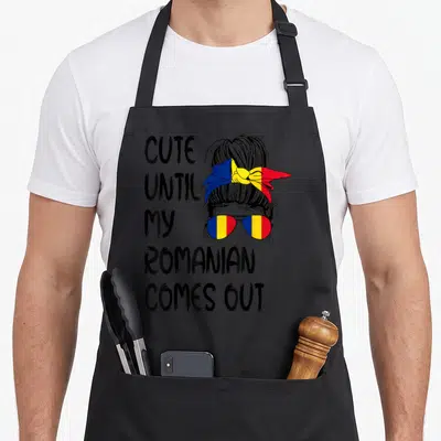 Come romanian apron - CUTE UNTIL MY ROMANIAN COMES OUT text next to woman with flag sunglasses and headband — shown on a black cotton canvas apron against a white background