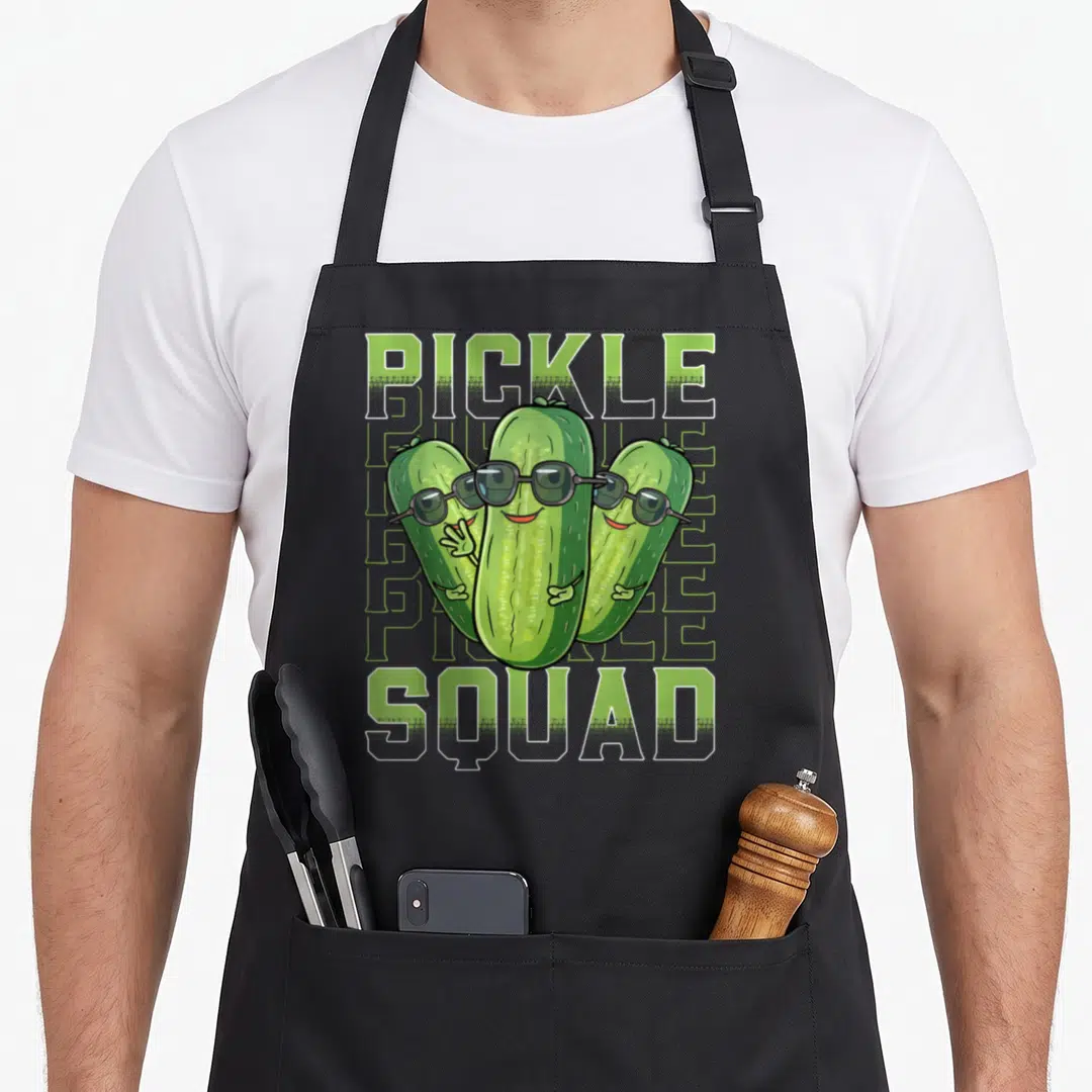 Squad pickle apron - Three cartoon pickles with sunglasses and green PICKLE SQUAD text — shown on a black cotton canvas apron against a white background