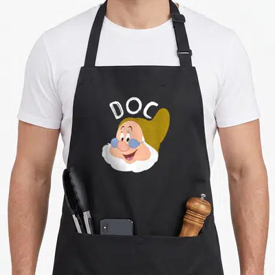 Doc snow white apron - Cartoon dwarf face with blue glasses and arched DOC text in olive and peach — shown on a black cotton canvas apron against a white background