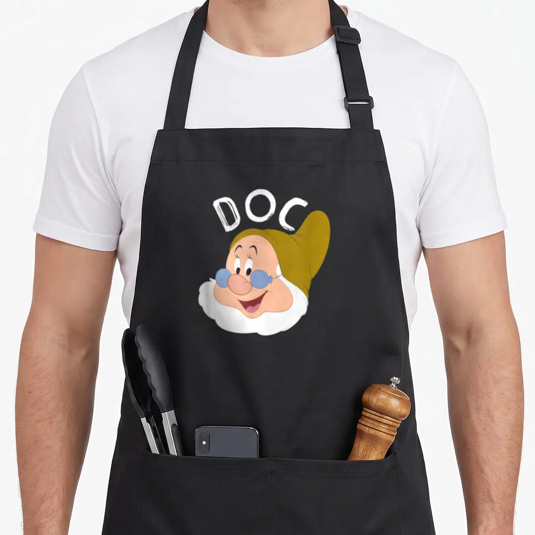 Doc snow white apron - Cartoon dwarf face with blue glasses and arched DOC text in olive and peach — shown on a black cotton canvas apron against a white background