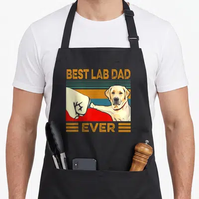 Lab owner father apron - Retro BEST LAB DAD EVER text above human hand fist bumping yellow labrador paw on striped — shown on a black cotton canvas apron against a white background