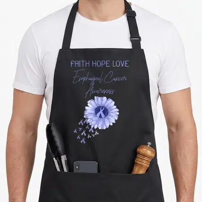 Esophageal ribbon apron - Purple flower with ribbon center and FAITH HOPE LOVE text stacked above — shown on a black cotton canvas apron against a white background