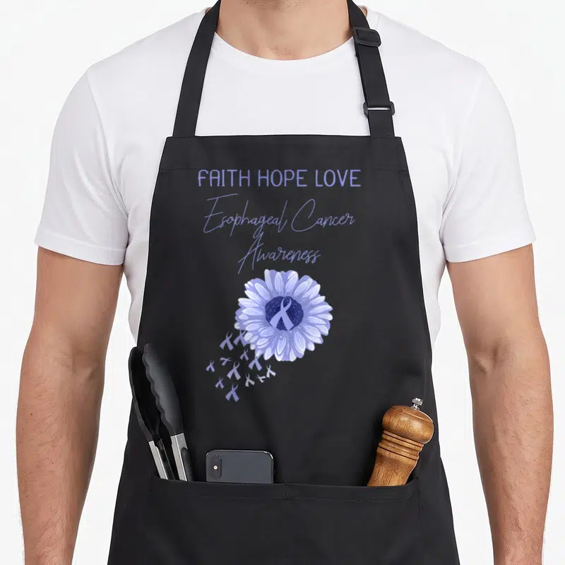 Esophageal ribbon apron - Purple flower with ribbon center and FAITH HOPE LOVE text stacked above — shown on a black cotton canvas apron against a white background
