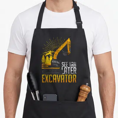 Yah excavator girl apron - Retro yellow excavator with See Yah Later text and mountain background — shown on a black cotton canvas apron against a white background