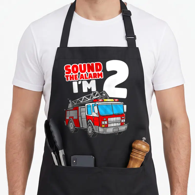 Fire firefighter truck apron - Red fire truck illustration with Sound the Alarm I'm 2 text in cartoon style — shown on a black cotton canvas apron against a white background