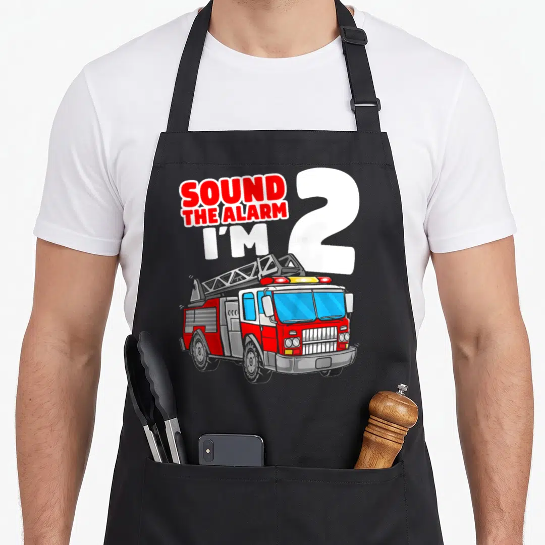 Fire firefighter truck apron - Red fire truck illustration with Sound the Alarm I'm 2 text in cartoon style — shown on a black cotton canvas apron against a white background