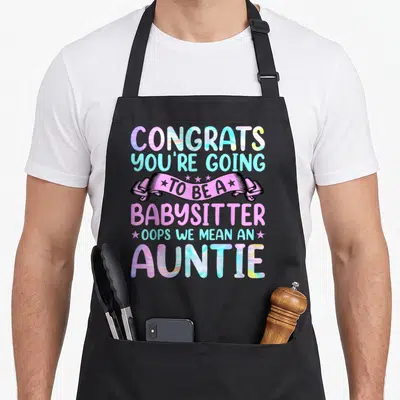 Aunt congrats apron - Holographic text CONGRATS YOU'RE GOING TO BE A BABYSITTER OOPS WE MEAN AN AUNTIE with pink ribbon — shown on a black cotton canvas apron against a white background