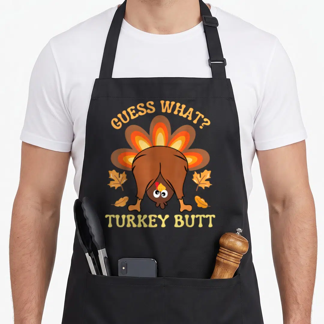 Guess turkey apron - Cartoon brown turkey rear view with arched orange text Guess What and straight yellow text Turkey — shown on a black cotton canvas apron against a white background