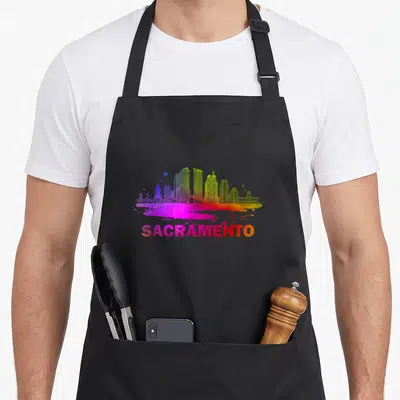 Sacramento colorful aprons - Rainbow skyline silhouette above paint splatter and gradient SACRAMENTO text in watercolor — shown on a black cotton canvas apron against a white background