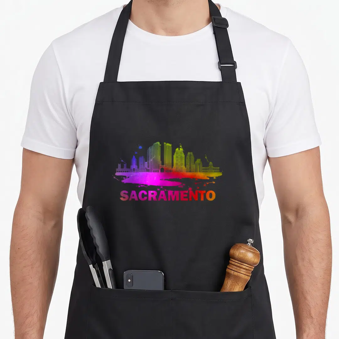 Sacramento colorful aprons - Rainbow skyline silhouette above paint splatter and gradient SACRAMENTO text in watercolor — shown on a black cotton canvas apron against a white background