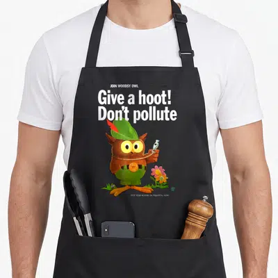 Woodsy give owl apron - Cartoon owl holding plastic bottle with Give a hoot Don't pollute text in green and brown — shown on a black cotton canvas apron against a white background