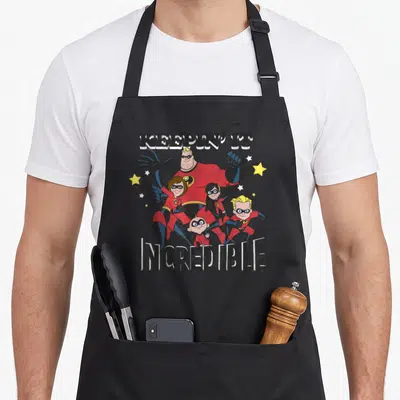 Keepin novelty apron - KEEPIN' IT INCREDIBLE text with cartoon superhero family and stars — shown on a black cotton canvas apron against a white background