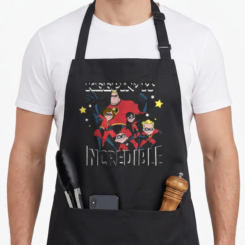 Keepin novelty apron - KEEPIN' IT INCREDIBLE text with cartoon superhero family and stars — shown on a black cotton canvas apron against a white background