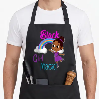 Melanin african print apron - Cartoon girl standing right of rainbow with stacked 'Black Girl MaGiC!' text in pink — shown on a black cotton canvas apron against a white background