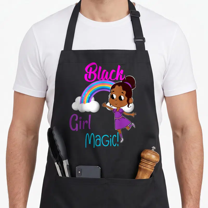 Melanin african print apron - Cartoon girl standing right of rainbow with stacked 'Black Girl MaGiC!' text in pink — shown on a black cotton canvas apron against a white background