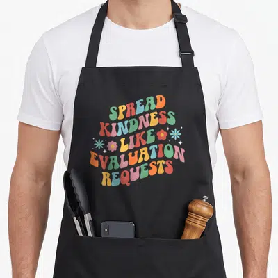 Spread psychologist apron - Wavy retro text reading SPREAD KINDNESS LIKE EVALUATION REQUESTS with small flowers — shown on a black cotton canvas apron against a white background