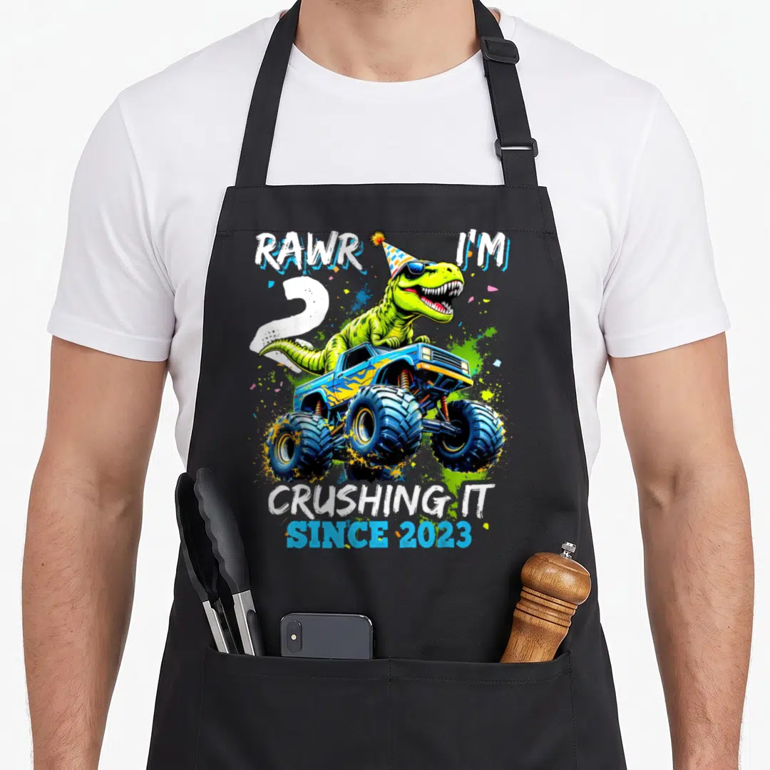 Dinosaur rawr truck apron - Green dinosaur in party hat on blue monster truck with RAWR I'M 2 text and confetti — shown on a black cotton canvas apron against a white background