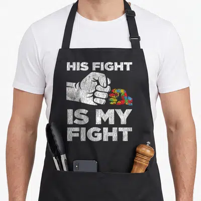 Awareness autism dad apron - Distressed white fist holding red blue yellow and green puzzle piece with text HIS FIGHT — shown on a black cotton canvas apron against a white background