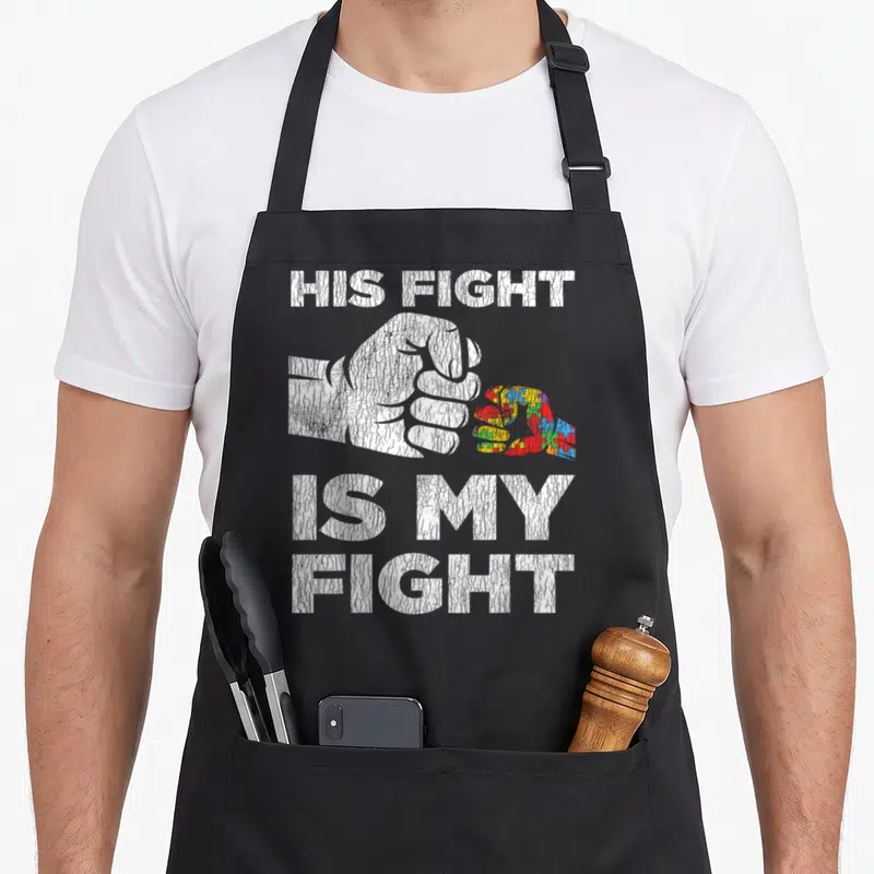 Awareness autism dad apron - Distressed white fist holding red blue yellow and green puzzle piece with text HIS FIGHT — shown on a black cotton canvas apron against a white background