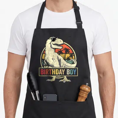 Dino family cooking aprons - Retro T-Rex in sunglasses with BIRTHDAY BOY banner over sunset circle — shown on a black cotton canvas apron against a white background