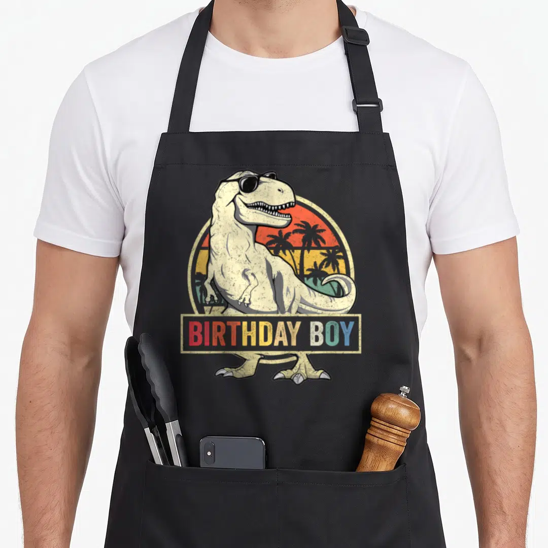 Dino family cooking aprons - Retro T-Rex in sunglasses with BIRTHDAY BOY banner over sunset circle — shown on a black cotton canvas apron against a white background