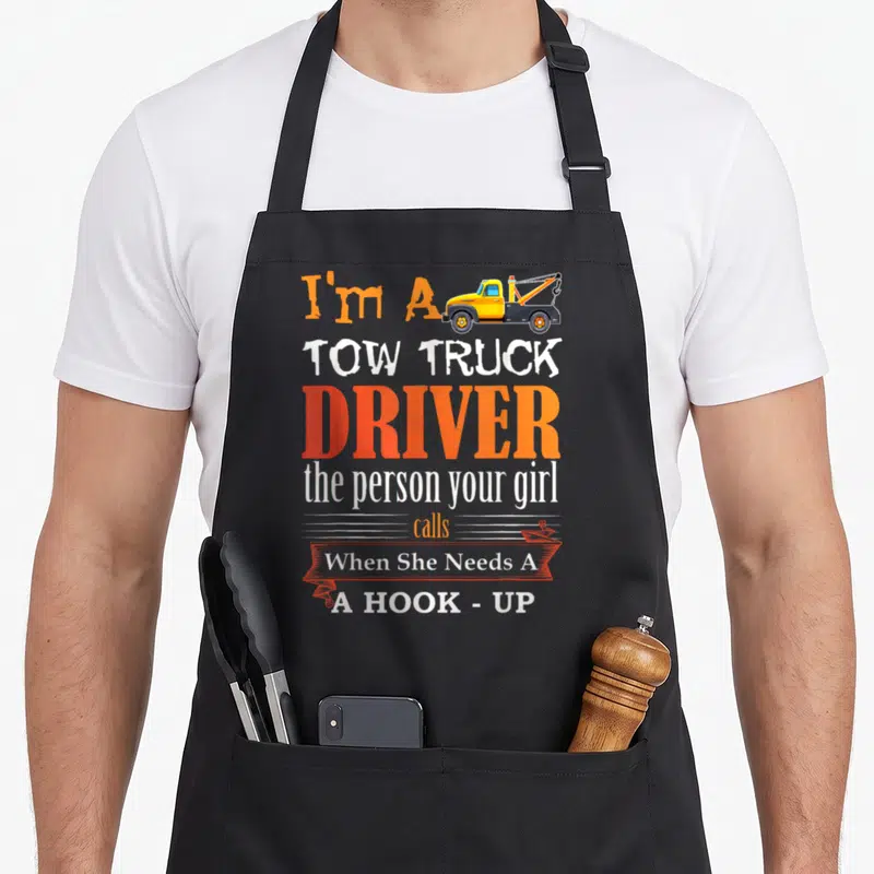 Wrecker truck apron - I'm A TOW TRUCK DRIVER text with yellow tow truck and orange banner reading When She Needs A HOOK — shown on a black cotton canvas apron against a white background