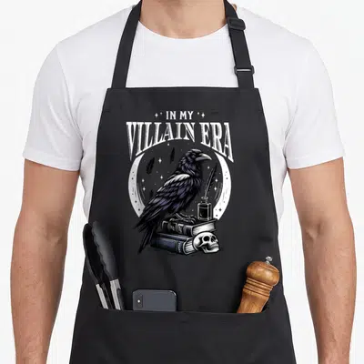 Villain bibliophile apron - Raven perched on books with skull inkwell and IN MY VILLAIN ERA text — shown on a black cotton canvas apron against a white background
