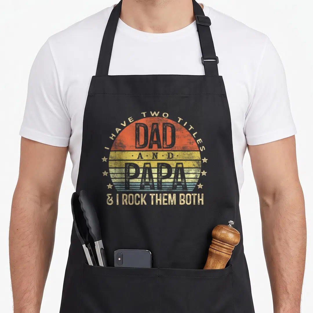 Title papa apron - Retro sun graphic with text I Have Two Titles Dad And Papa I Rock Them Both surrounded by stars — shown on a black cotton canvas apron against a white background