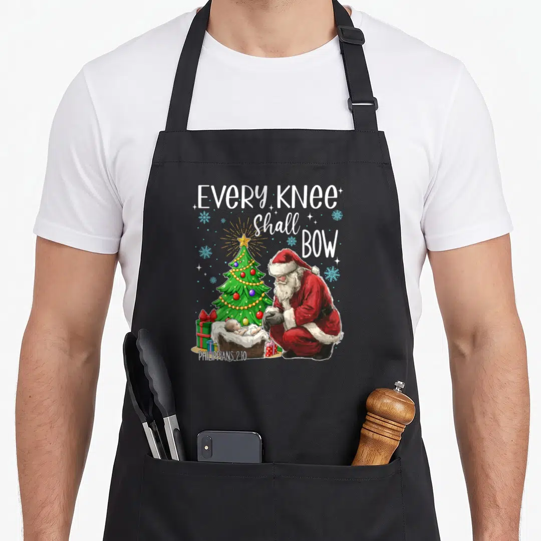 Knee santa apron - Santa kneeling before baby Jesus with Every Knee Shall Bow text and Christmas tree — shown on a black cotton canvas apron against a white background