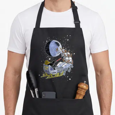 Vader open apron - Darth Vader in sleigh pulled by three AT-AT walkers with antlers near Death Star — shown on a black cotton canvas apron against a white background