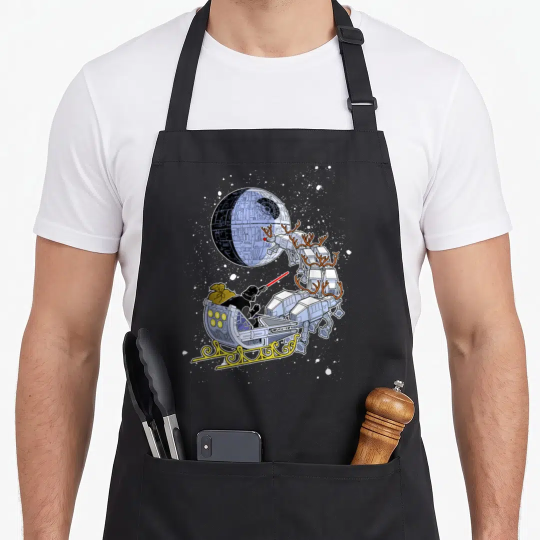 Vader open apron - Darth Vader in sleigh pulled by three AT-AT walkers with antlers near Death Star — shown on a black cotton canvas apron against a white background
