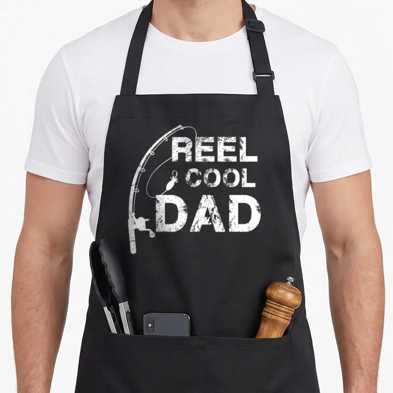 Reel daddy aprons - White distressed fishing rod and lure beside stacked REEL COOL DAD text — shown on a black cotton canvas apron against a white background