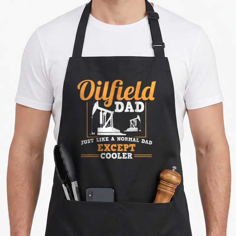 Oilfield rig dad apron - Two white pumpjacks in orange frame with distressed Oilfield Dad text — shown on a black cotton canvas apron against a white background