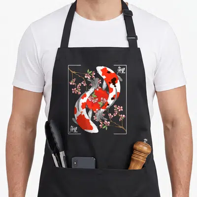 Carp cherry apron - Two koi fish circling red sun with pink blossoms and kanji characters in white frame — shown on a black cotton canvas apron against a white background