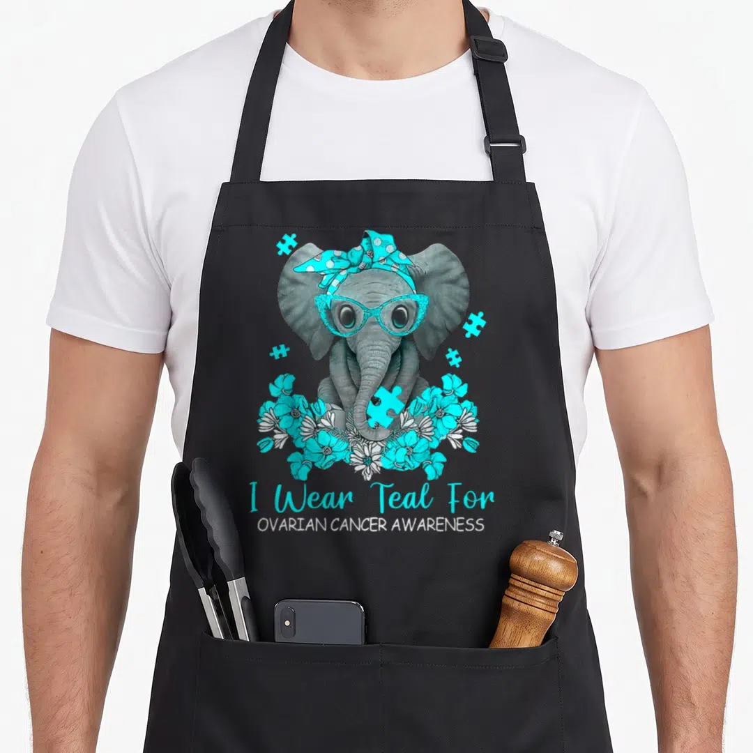 Elephant cancer girl apron - Grey cartoon elephant with teal glasses bandana and puzzle pieces holding flower cluster — shown on a black cotton canvas apron against a white background
