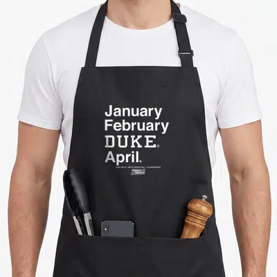 Basketball march madness apron - White stacked text reading January February DUKE April with NCAA logo — shown on a black cotton canvas apron against a white background