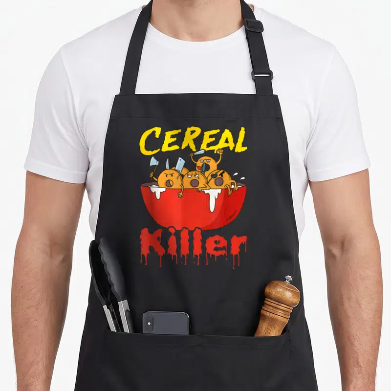 Serial cereal apron - Red bowl with angry orange cereal pieces holding knives and axes under yellow text — shown on a black cotton canvas apron against a white background