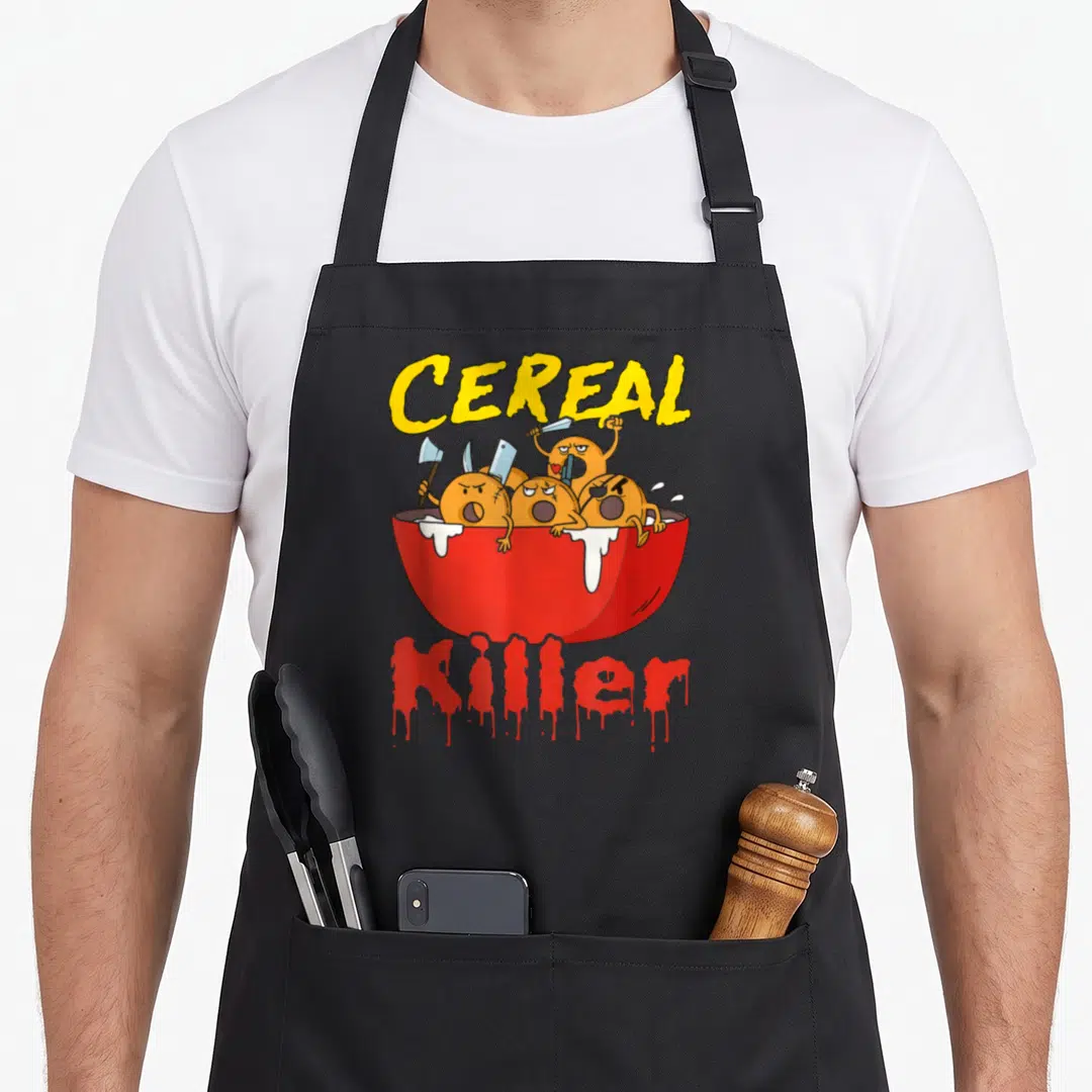 Serial cereal apron - Red bowl with angry orange cereal pieces holding knives and axes under yellow text — shown on a black cotton canvas apron against a white background