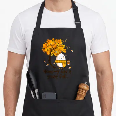 Humpty cute fall aprons - Egg character in scarf holding coffee under orange tree with Humpty had a great fall text — shown on a black cotton canvas apron against a white background