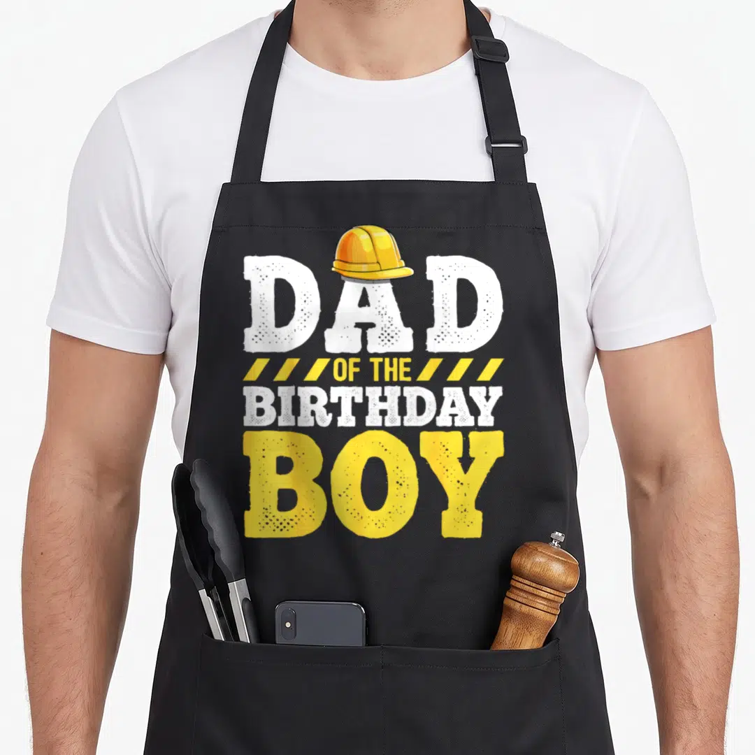 Birthday hat construction apron - Stacked white and yellow text reading Dad of the Birthday Boy with a yellow hard hat — shown on a black cotton canvas apron against a white background